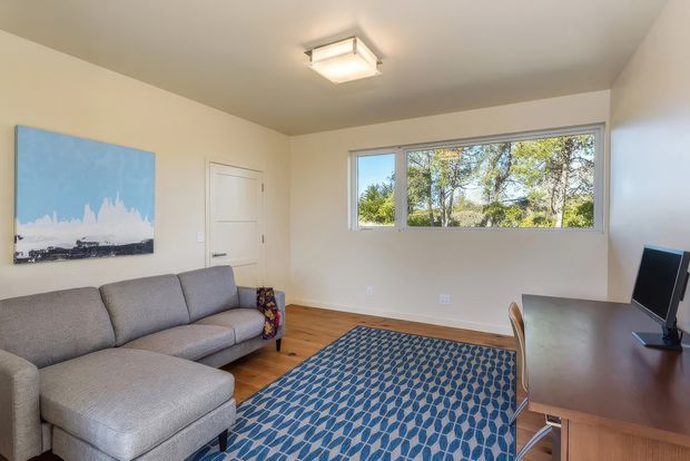 A compact, bright living room or study with a grey sectional sofa, patterned blue area rug, timber flooring, white walls, a simple ceiling light, and a dark wood desk with monitor. A wide horizontal window provides views of trees and daylight.