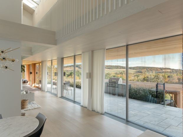 Bright contemporary open-plan living and dining area with a double-height void, extensive floor-to-ceiling glazing, pale timber flooring, and a white balustrade overlooking the room. The space opens onto a tiled terrace with an outdoor grill, while rolling landscape views are visible beyond the glazing. A sculptural chandelier, dining table, and partial seating area add residential character.