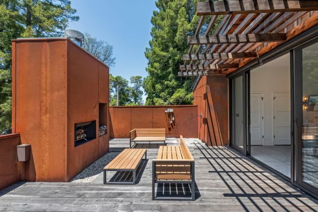 A contemporary outdoor terrace with a timber deck, built-in rust-coloured rendered fireplace or feature wall, timber pergola, and two wooden benches with black metal frames. Large glazed sliding doors connect the terrace to the interior, while mature trees surround the space.