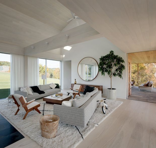Bright, spacious contemporary living room with extensive floor-to-ceiling glazing, pale timber flooring and ceiling, white walls, exposed ceiling beams, and a large textured neutral rug. Two striped sofas and mid-century-inspired timber lounge chairs surround a low rectangular coffee table. A round wall mirror, sculptural planter, and indoor tree add warmth. An open doorway leads to a secondary furnished room or terrace area, while landscaped grounds are visible through the glazing.