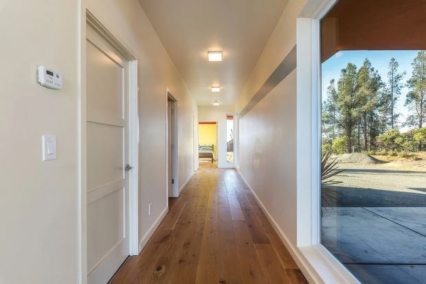 Long, bright residential hallway with wide-plank timber flooring, pale painted walls, white paneled interior doors and trim, recessed ceiling lights, and a large floor-to-ceiling glazed opening at the right overlooking a driveway and trees. A bedroom or living area is visible at the far end.