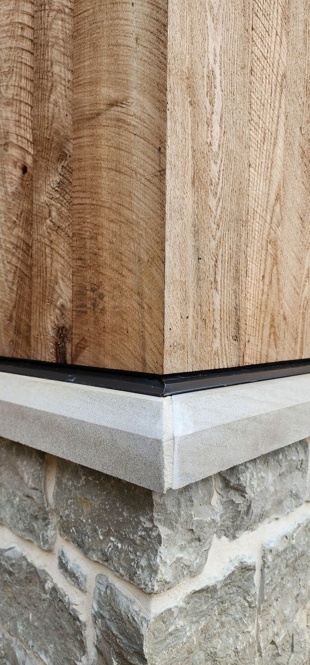 Close-up detail of an exterior building corner showing rough-sawn timber cladding above a dark flashing or trim, a pale concrete or stone projecting ledge, and irregular grey natural stone masonry below. The image focuses on the junction between timber, foundation trim, and masonry construction.