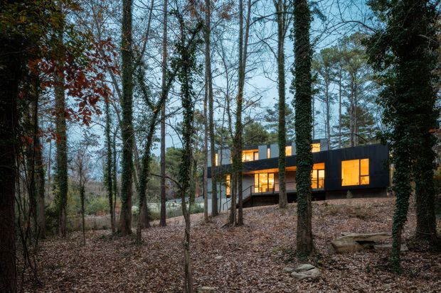 A modern, dark-clad multi-level house nestled among mature woodland. Large warm-lit windows punctuate the rectilinear facade, with a gently sloping wooded site and exposed natural landscape surrounding the building.