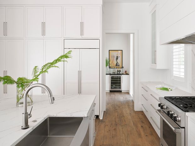 Bright, spacious white galley-style kitchen with extensive shaker-style cabinetry, a marble-look island and benchtops, a large stainless-steel undermount sink with gooseneck tap, and a stainless-steel range. White horizontal wall panelling and plantation shutters frame the room, while a timber floor leads to a small adjoining beverage or bar area with display shelving and a wine fridge.