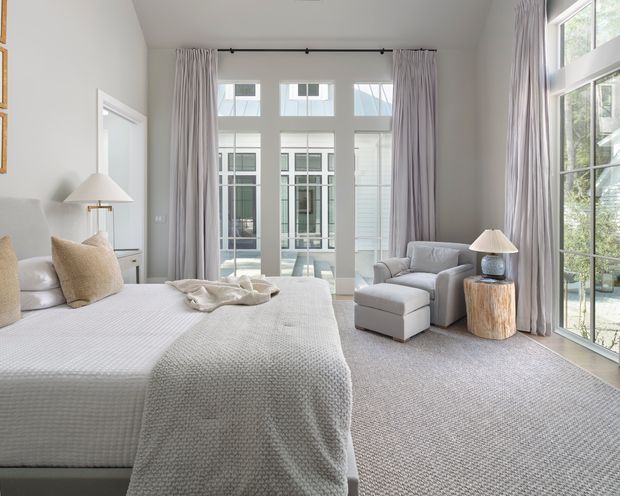 Bright, spacious bedroom with a soft neutral palette, a large upholstered bed, pale textured bedding, floor-to-ceiling curtains, and extensive glazing overlooking an exterior courtyard or adjacent structure. A seating nook with armchairs and a stump-style side table occupies the right side of the room. The space has a refined, calm contemporary feel and a high ceiling.