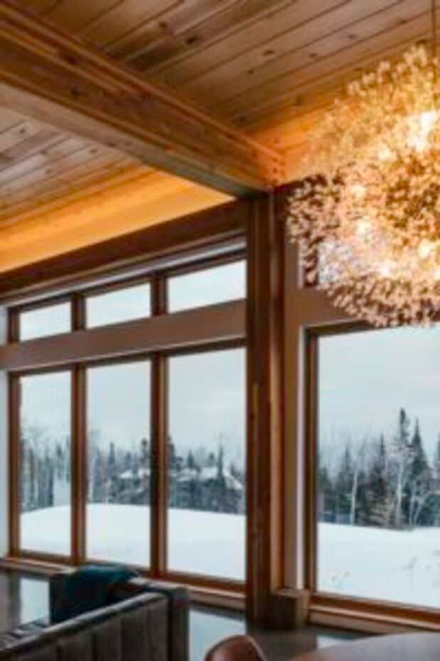 Interior view of a warm, rustic timber-lined room with exposed wood ceiling beams, substantial timber window frames, and expansive glazing overlooking a snowy forested landscape. A sectional sofa and part of a table are visible in the foreground, with a large illuminated branching decorative light fixture near the upper right.