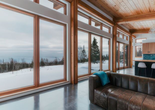 A bright contemporary living area with expansive floor-to-ceiling timber-framed glazing overlooking a snowy landscape. The room features a polished dark concrete floor, timber-lined ceiling, exposed timber posts, and a dark brown leather sectional sofa with a teal throw. A compact kitchen or bar area with timber cabinetry and a blue-green tiled splashback is visible at the rear.
