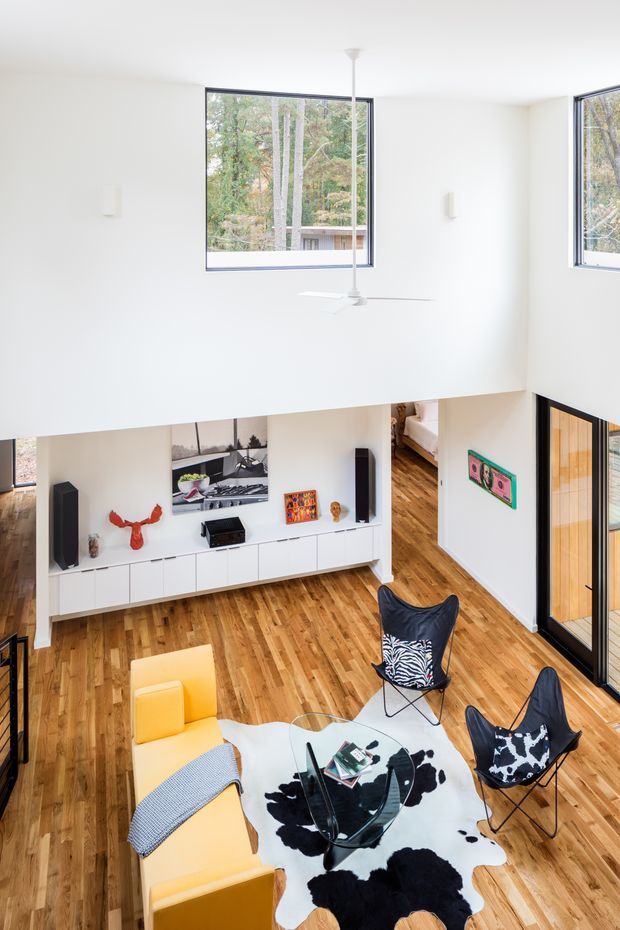 Bright double-height living room with a mezzanine-like upper wall and large corner windows overlooking trees. The room has white walls and ceiling, warm timber flooring, a built-in white media cabinet, ceiling fan, and a seating arrangement with yellow upholstered chairs, black leather-and-metal butterfly chairs, a glass coffee table, and a black-and-white cowhide rug. A glazed timber-framed door or partition is visible at right, with a bedroom partially visible beyond the open passage.