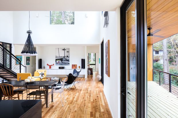 Bright, open-plan contemporary living and dining space with a double-height ceiling, exposed timber flooring, black metal stair and balustrade, large glazed doors, and a timber-lined covered deck visible outside. The room includes a dining table, lounge seating, artwork, and a sculptural pendant light. A mezzanine or upper-level void is suggested by the stair and high ceiling.