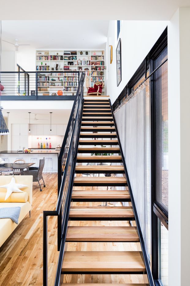 A bright, double-height contemporary interior centred on an open staircase with light timber treads and slim black metal railings. The stair rises beside tall black-framed glazing and overlooks an open-plan kitchen and dining area with timber flooring. A mezzanine-level library with extensive built-in white shelving and books is visible beyond the landing. White walls, abundant daylight, and the open stair construction create an airy, modern residential atmosphere.