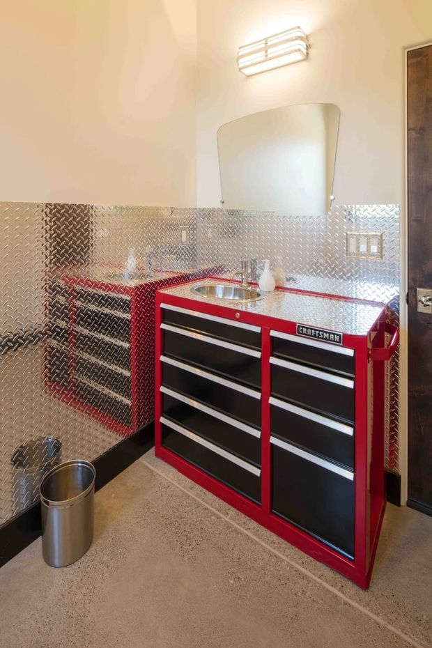 Compact industrial-style utility bathroom or washroom featuring a red-and-black Craftsman tool-chest vanity with integrated stainless-steel sink, diamond-plate metal wall cladding, a rounded rectangular mirror, exposed-style wall light, and polished concrete flooring. A dark timber door is visible at the right.
