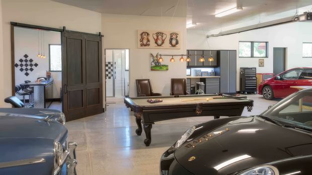 A spacious, bright garage or showroom with polished concrete flooring, exposed ceiling services, dark industrial-style sliding doors, automotive workbenches and tool storage, and several classic and modern cars surrounding a central billiards table. Decorative automotive-themed artwork and pendant lights add character.