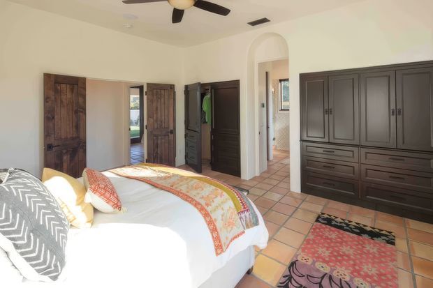 A bright bedroom with a large bed in the foreground, patterned bedding and pillows, dark built-in wardrobe cabinetry, and multiple dark timber doors. The room has pale walls, terracotta-toned tiled flooring, an arched opening, and a ceiling fan. A bathroom or adjoining tiled space is visible through the arched doorway.