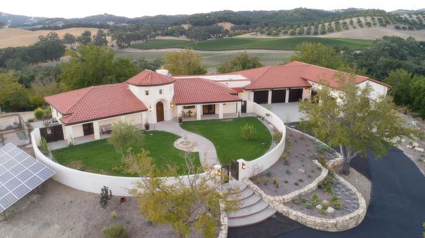Aerial view of a large Mediterranean-style rural residence with white stucco walls, multiple red terracotta-tiled roofs, arched openings, and a curved white perimeter wall. The landscaped grounds include a circular lawn courtyard, curved stone-edged garden beds, a paved driveway, and visible solar panels. Rolling agricultural hills and vineyard-like planting surround the property.