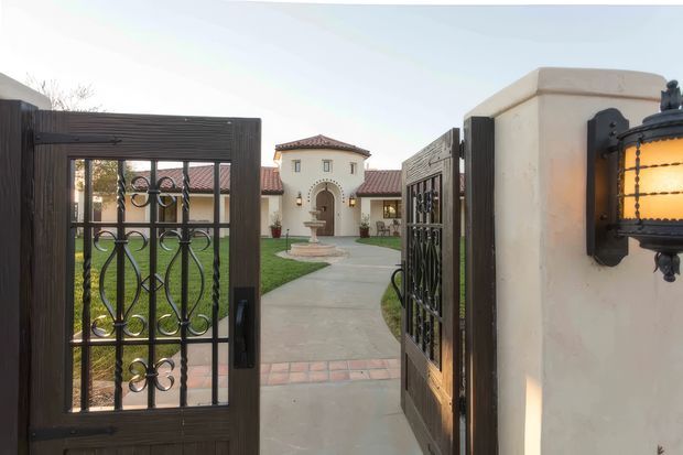 View through open wrought-iron pedestrian gates into a formal courtyard entry. The property features cream stucco walls, dark timber-toned gate framing, decorative black metalwork, terracotta roof tiles, arched detailing, a central fountain, and a paved walkway leading to a Mediterranean-style house. A wall-mounted lantern is visible in the foreground.