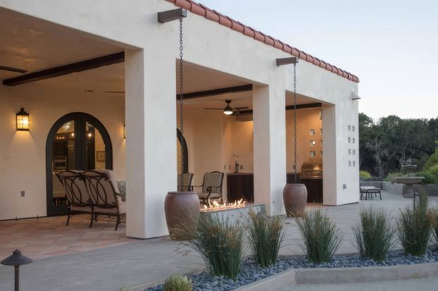 Exterior view of a Mediterranean-inspired covered patio with thick white stucco columns, terracotta roof-edge tiles, arched timber-framed doors, hanging chain-supported planters, and a linear fire feature. A dining area and lounge furnishings sit beneath the covered verandah, with gravel landscaping and ornamental grasses in the foreground.
