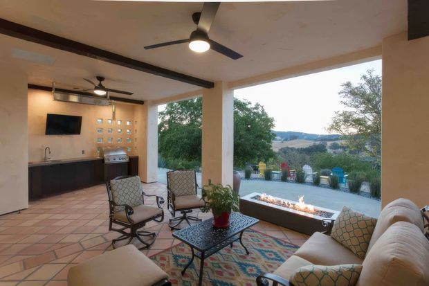 A covered outdoor living and entertaining area with a built-in kitchen, tiled floor, ceiling fans, cushioned seating, patterned rug, and a long linear fire feature. Large open sides frame a landscaped outlook toward rolling countryside.
