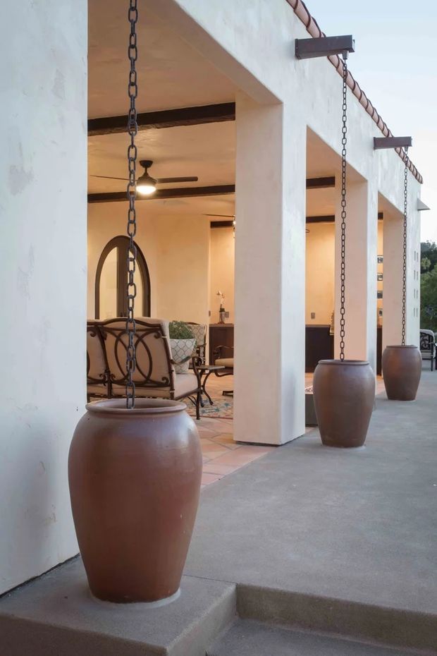 A covered outdoor terrace with large white stucco columns and exposed dark timber beams, featuring a row of suspended ceramic rainwater collection pots attached to hanging chains. The terrace opens into a warm, Spanish-inspired living or dining area with arched openings, terracotta floor tiles, wrought-iron furniture, ceiling fans, and soft ambient lighting.