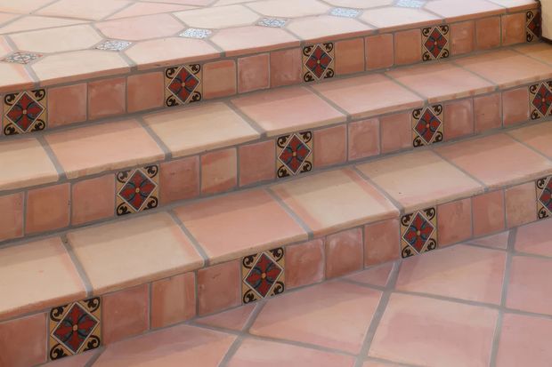 Close-up view of tiled architectural steps finished in terracotta and pale pink tiles. The risers feature regularly spaced decorative diamond-pattern accent tiles with dark blue, red, cream, and black motifs, while the surrounding floor and stair surfaces use irregular geometric tile layouts with contrasting grout lines.