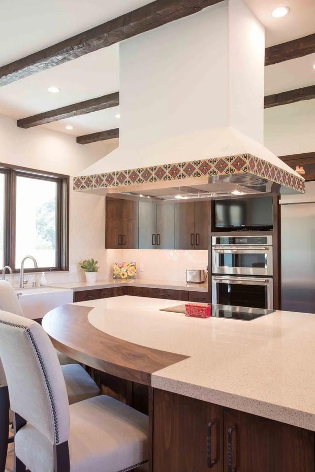 Bright, spacious kitchen with a large curved peninsula island featuring a light speckled stone benchtop and integrated cooktop. Dark timber cabinetry, stainless-steel appliances, white walls, exposed dark timber ceiling beams, and a prominent custom rangehood surround with colourful patterned tile trim define the room. A window, sink, dining-height seating, and built-in double oven are visible.