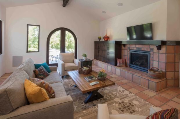 Warm, relaxed living room with a large upholstered sectional sofa, armchairs, timber coffee table and patterned rug arranged around a substantial tiled fireplace. The room has pale plaster walls, terracotta-toned floor and fireplace tiles, dark timber built-in cabinetry, an arched glazed door, and an exposed ceiling beam. A wall-mounted television sits above the fireplace.