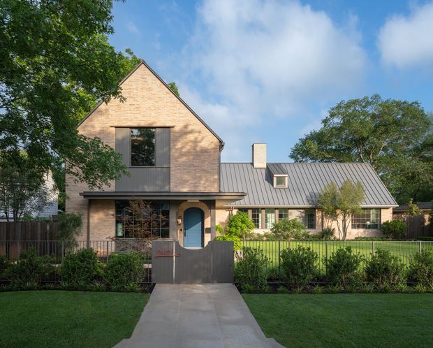 Front exterior view of a substantial residential house with a steep gabled brick volume connected to a lower wing with a standing-seam metal roof. The facade combines warm pale brick, dark-framed glazing, a projecting entry canopy, and a distinctive blue arched front door. A straight concrete path leads through a landscaped front garden and low fence. Mature trees and bright sky surround the property.