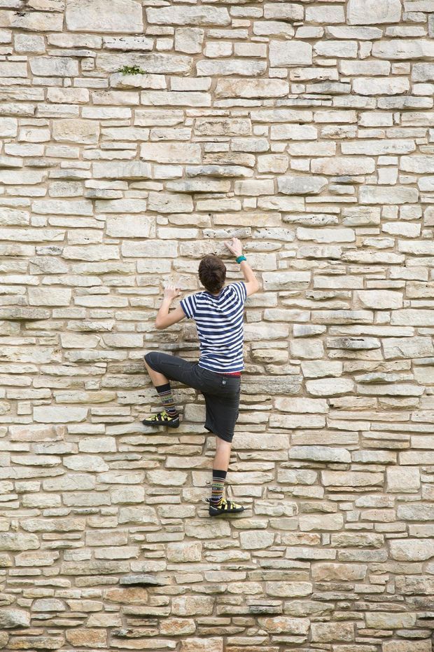 A person wearing climbing shoes and a striped shirt is scaling a tall, irregularly coursed pale stone masonry wall. The image is dominated by the textured exposed stone surface, with no clearly visible building interior or broader architectural context.