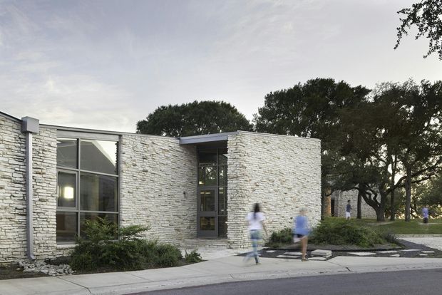 Low, single-storey contemporary building with a predominantly horizontal composition, clad in pale stacked stone masonry. Large dark-framed glazed openings and a recessed glazed entry sit beneath a shallow projecting roof, with mature trees and landscaped planting around the paved approach.