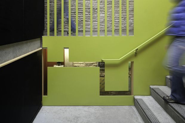 Interior stairwell with bright lime-green painted walls and balustrade, exposed textured stonework visible behind vertical metal railings and through wall openings, dark wall panels on the left, and grey carpeted concrete stairs rising on the right. A person is partially visible on the stairs. The space has a bold, graphic, contemporary institutional or commercial character.