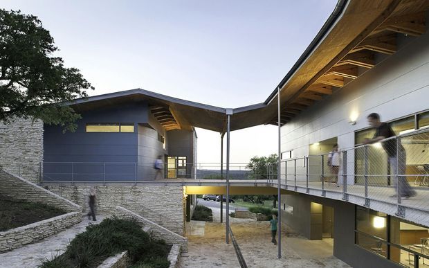 Exterior architectural view of a contemporary multi-level building complex arranged around a landscaped courtyard and elevated pedestrian bridge. The composition features dark metal-clad volumes, pale horizontal wall cladding, extensive exposed timber soffits and rafters, stone retaining walls, open steel railings, and covered circulation spaces. The building has strongly projecting pitched roof planes and an integrated lower-level courtyard or entry area.