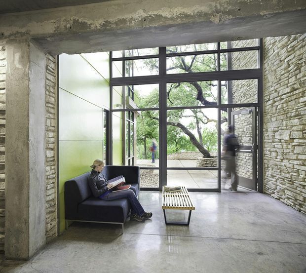 A modern, industrial-style reading and circulation area framed by exposed concrete beams and columns, rough stacked-stone walls, polished concrete flooring, and large black-framed floor-to-ceiling glazing. A dark lounge chair and low slatted coffee table sit beside the entry, with a landscaped courtyard and mature tree visible beyond the glass.