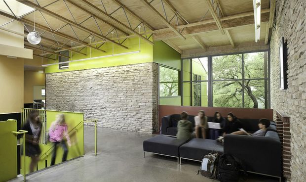 A bright, informal multi-level interior lounge or common area with exposed timber roof structure and services, polished concrete flooring, extensive rough stone masonry walls, lime-green painted walls and partitions, and large black-framed windows overlooking trees. Dark upholstered modular seating forms a social gathering area, while a stair or sunken-level transition is visible at left. The space has an adaptive industrial character with strong natural materials and high ceilings.
