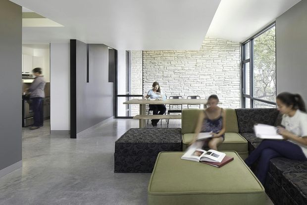 Open-plan contemporary interior showing a lounge and study area, with upholstered modular seating in the foreground, a long dining or work table near full-height glazing, dark built-in joinery, polished concrete flooring, and a white exposed brick feature wall. The space is bright and connected to a kitchen or service area at left.