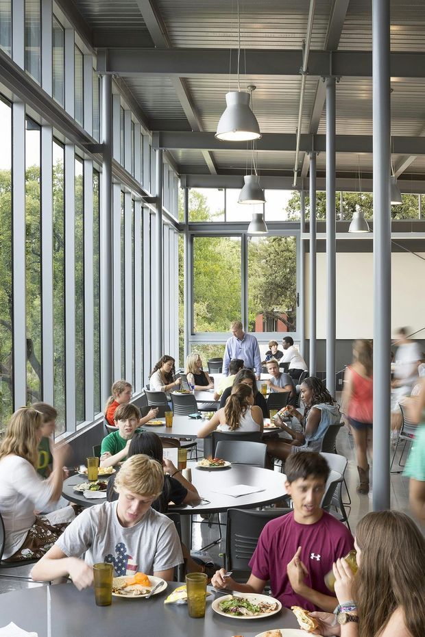 A bright, industrial-style communal dining area or cafeteria with many people seated at round tables. The space features extensive floor-to-ceiling glazing, exposed grey steel columns and beams, a corrugated metal ceiling, suspended cone pendant lights, and dark tables and chairs. Trees are visible outside through the windows.