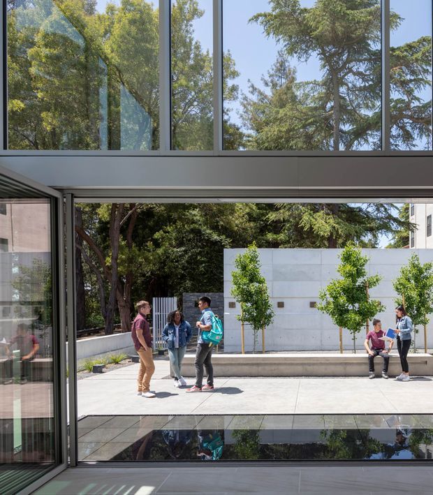 A contemporary glazed interior or circulation space opens onto a landscaped courtyard through large sliding glass doors. Floor-to-ceiling windows and clerestory glazing frame mature trees, while a shallow reflecting pool or dark water feature sits outside. Several people gather along a concrete bench beside a pale rendered or concrete wall with regularly spaced small trees. The space is bright, clean, and strongly connected to the outdoors.