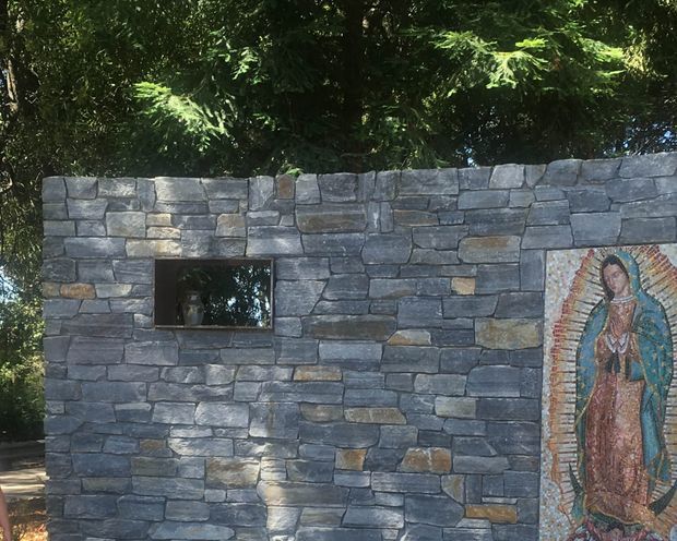 Outdoor stone masonry wall built from irregular stacked grey and blue-grey stone, with a small dark rectangular inset or opening and a colourful religious mosaic panel mounted at the right. Dense leafy vegetation rises behind the wall, creating a shaded garden-like setting.