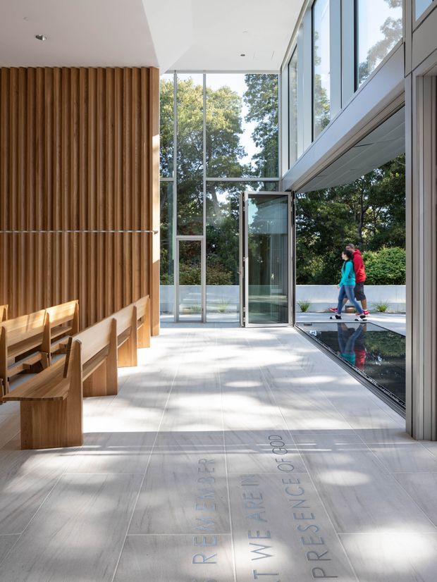 Bright contemporary interior or institutional lobby with full-height glazing, pale tiled flooring, a sculptural vertical timber-slat feature wall, integrated timber bench seating, and a long shallow reflecting pool along the glazed edge. People are visible walking outside, with landscaped greenery beyond. The space has a refined, airy, gallery-like atmosphere and strong indoor–outdoor connection.