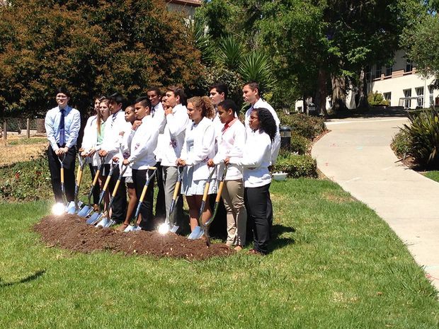 A group of people in white shirts pose outdoors with shovels beside a freshly dug patch of soil, suggesting a ceremonial groundbreaking or landscaping event. The scene is set on a landscaped grassy campus or property with mature trees, planted garden beds, and a paved walkway leading toward a building.