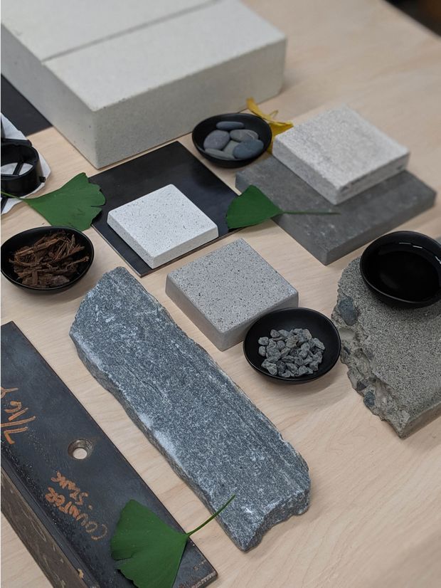 A top-down display of architectural and landscape material samples arranged on a light timber surface. The collection includes rough and honed grey stone slabs, speckled concrete or stone tiles, a dark slate-like board, bowls of small stones and organic fragments, and green leaf accents. The image appears to be a material selection or design sample presentation.