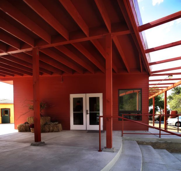 Exterior view of a red-painted timber building entrance beneath a deep exposed-beam canopy. The covered entry has concrete paving, accessible ramps and red metal handrails, white-framed double doors, a large window, and a translucent roof section extending along the side.