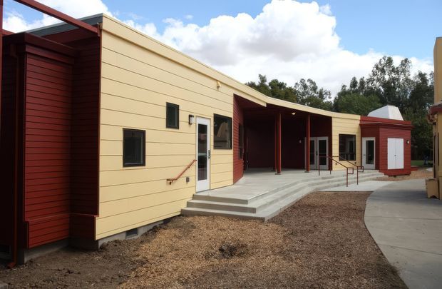Single-storey contemporary building exterior with a long low-slung form, cream horizontal cladding, deep red timber-clad projections and covered entry areas. Broad concrete steps lead to glazed entrance doors, with accessible handrails and a curved concrete pathway through a sparse landscaped forecourt.