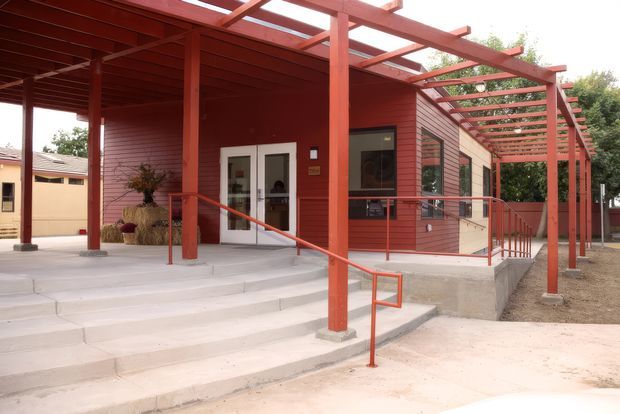 Exterior view of a low-rise red-clad building entrance with a broad concrete stairway, accessible ramp, red metal handrails, and a large timber pergola extending over the approach. Glazed double doors and windows reveal a modest interior, while adjacent buildings and landscaping sit in the background.
