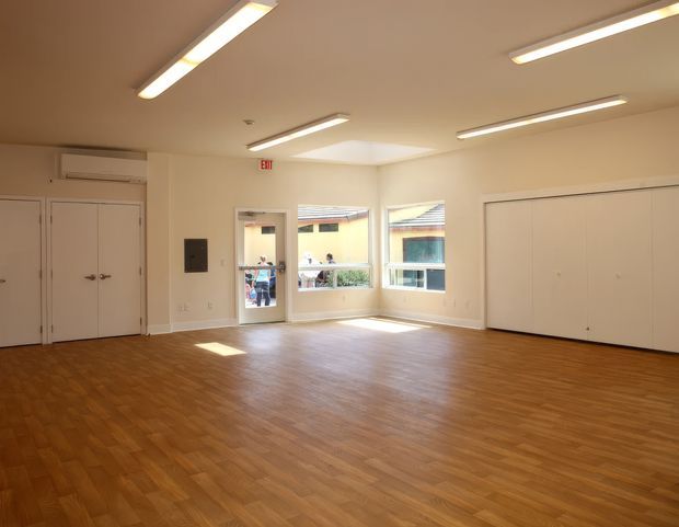 A bright, empty multi-purpose interior room with warm wood-pattern flooring, cream walls, white double doors and large built-in cupboards. Several linear fluorescent ceiling lights provide even illumination, while windows and a glazed door bring in daylight. The room appears suitable for a studio, classroom, office, or community/commercial use.