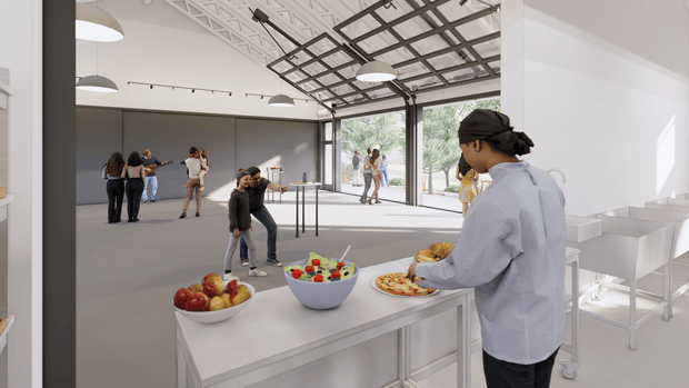 Bright, spacious contemporary multi-purpose interior with a large open floor area, exposed decorative ceiling structure, extensive glazed garage-style doors, and a serving island in the foreground. A person is preparing food at the island, while groups of people gather and move through the open space. White walls, grey flooring, linear pendant lighting, and a clean utilitarian fit-out create a flexible event or commercial space.