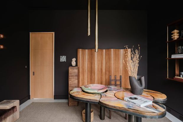 Dark, refined interior featuring a sculptural organic-shaped dining table with layered stone or timber-look surfaces, surrounded by dark cylindrical legs. A warm timber door and matching vertical timber wall panelling contrast with matte black walls and built-in shelving. Decorative objects, books, vessels, and pendant lighting create a sophisticated dining or entertaining setting.