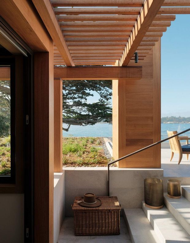 A refined coastal residential entry or transitional outdoor living space framed by substantial warm timber beams and a slatted pergola roof. Concrete steps and low walls lead toward a waterfront terrace, with a woven basket and coffee cup in the foreground. A large opening reveals landscaped planting, water, and a tree beyond, while outdoor dining furniture is visible to the right. The composition emphasises indoor-outdoor connection, natural materials, strong timber structure, and abundant daylight.