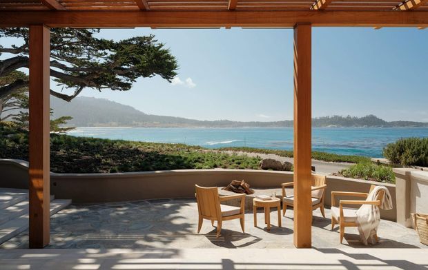 A covered beachfront outdoor terrace with a timber pergola, substantial timber posts and a patterned timber ceiling. The terrace is furnished with wooden lounge chairs and a small table, opening directly toward a bright turquoise bay, sandy shoreline, coastal vegetation, and distant hills. Strong indoor-outdoor connection with warm natural materials and expansive coastal views.