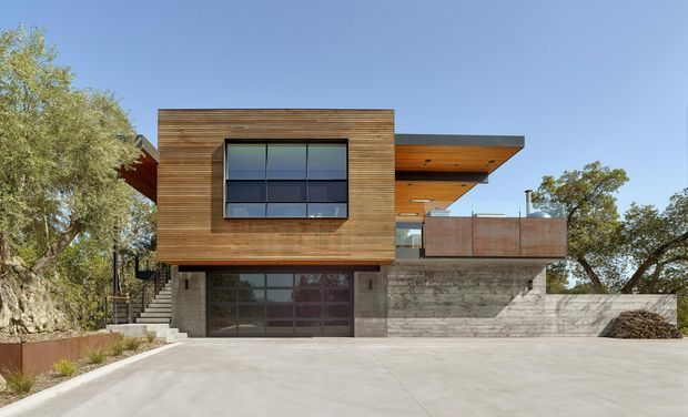 Exterior view of a contemporary multi-level residence on a sloped site. The house features a projecting box clad in horizontal timber, extensive dark-framed glazing, exposed concrete retaining and garage walls, and broad cantilevered roof and deck elements. A large concrete driveway occupies the foreground, with mature trees around the property.