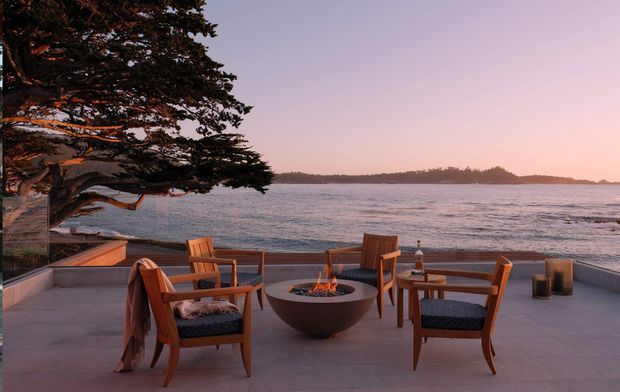 A waterfront outdoor terrace at sunset, furnished with timber armchairs and a low circular fire pit. A large coastal tree frames the left side, while a low balustrade overlooks calm water and a distant wooded shoreline. The space has a warm, relaxed, resort-like atmosphere with muted pink, blue-grey, timber and concrete tones.