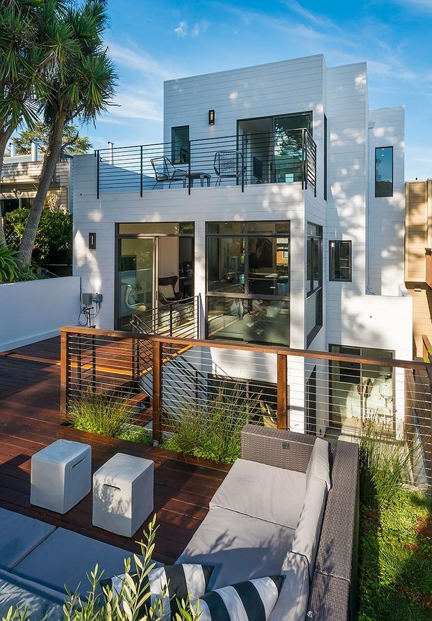 A contemporary multi-storey residence viewed from an elevated outdoor terrace. The white horizontally clad facade has large glazed openings, projecting balconies, dark metal railings, and a rooftop or upper-level timber deck furnished with an outdoor sectional sofa and cube stools. Landscaping borders the deck, while a palm tree and blue sky frame the scene.